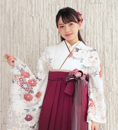 白地に多色の花柄が描かれた二尺袖と、エンジ色の袴を着て、可憐に微笑む女性。卒業式の袴スタイルで、腰元のリボンと花の飾りがアクセント。