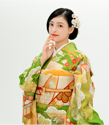 鮮やかな黄緑色に松や菊、梅などの古典的な柄が描かれた振袖を着て、口元に手を添えて微笑む女性。金色の帯と赤い帯締めがアクセント。